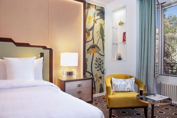 A hotel room with a bed, a nightstand, a lamp, a chair, a small table with a drink, a window with curtains, and a wall with a floral mural. - Hotel Bristol (Geneva)