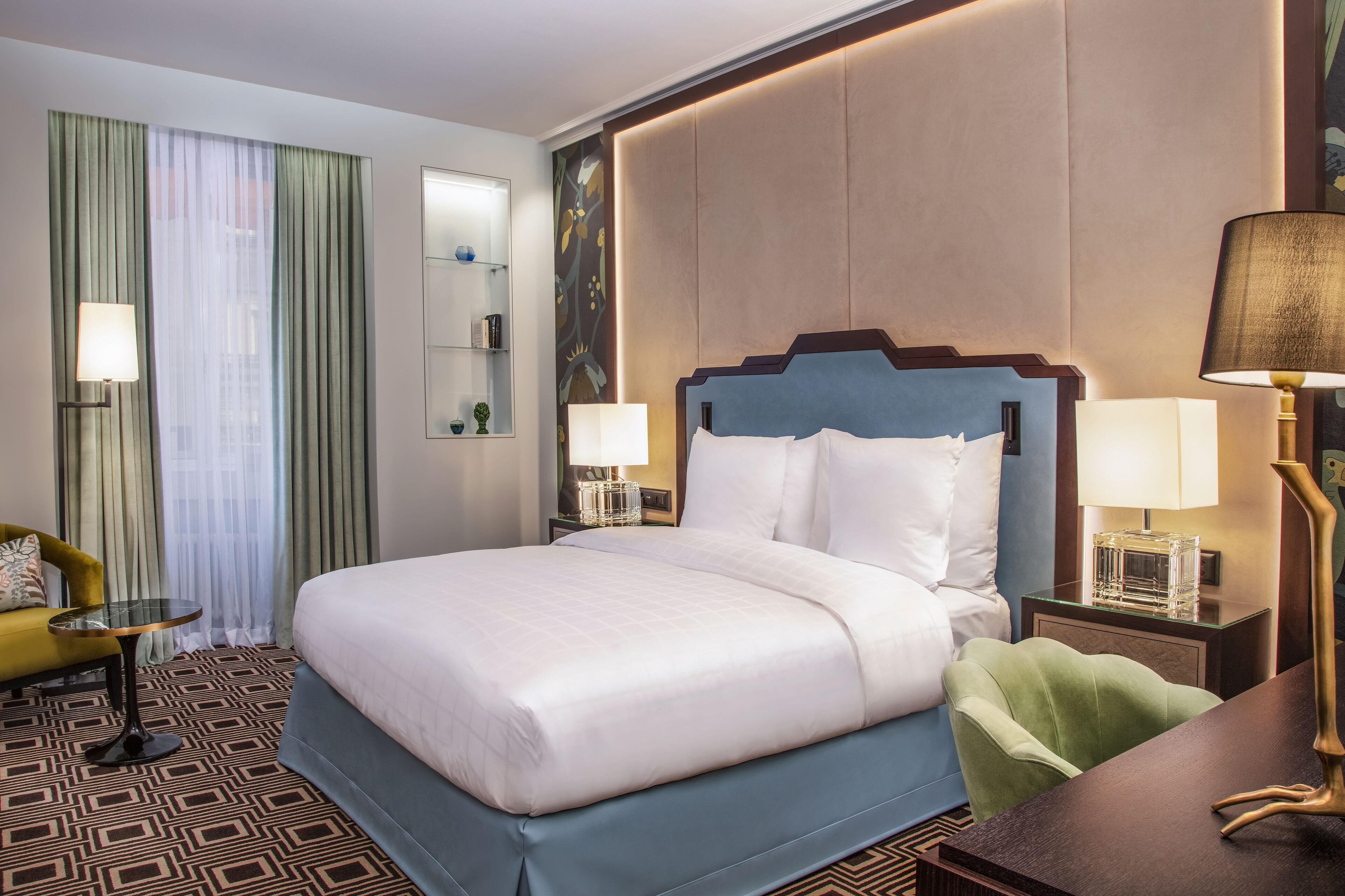 Habitación de hotel con una cama grande, dos mesitas de noche con lámparas, un sillón verde, una mesita redonda pequeña y una alfombra estampada.