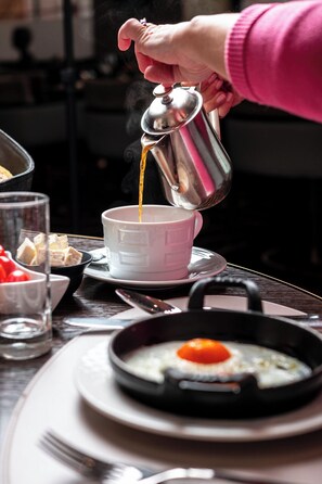 A hand pouring a hot beverage into a cup, with a plate of food and a frying pan with an egg on a table. - Hotel Bristol (Geneva)