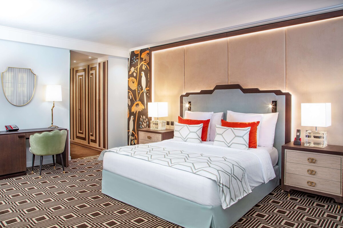 A hotel room with a large bed, two bedside tables, a desk with a chair, and a patterned carpet.