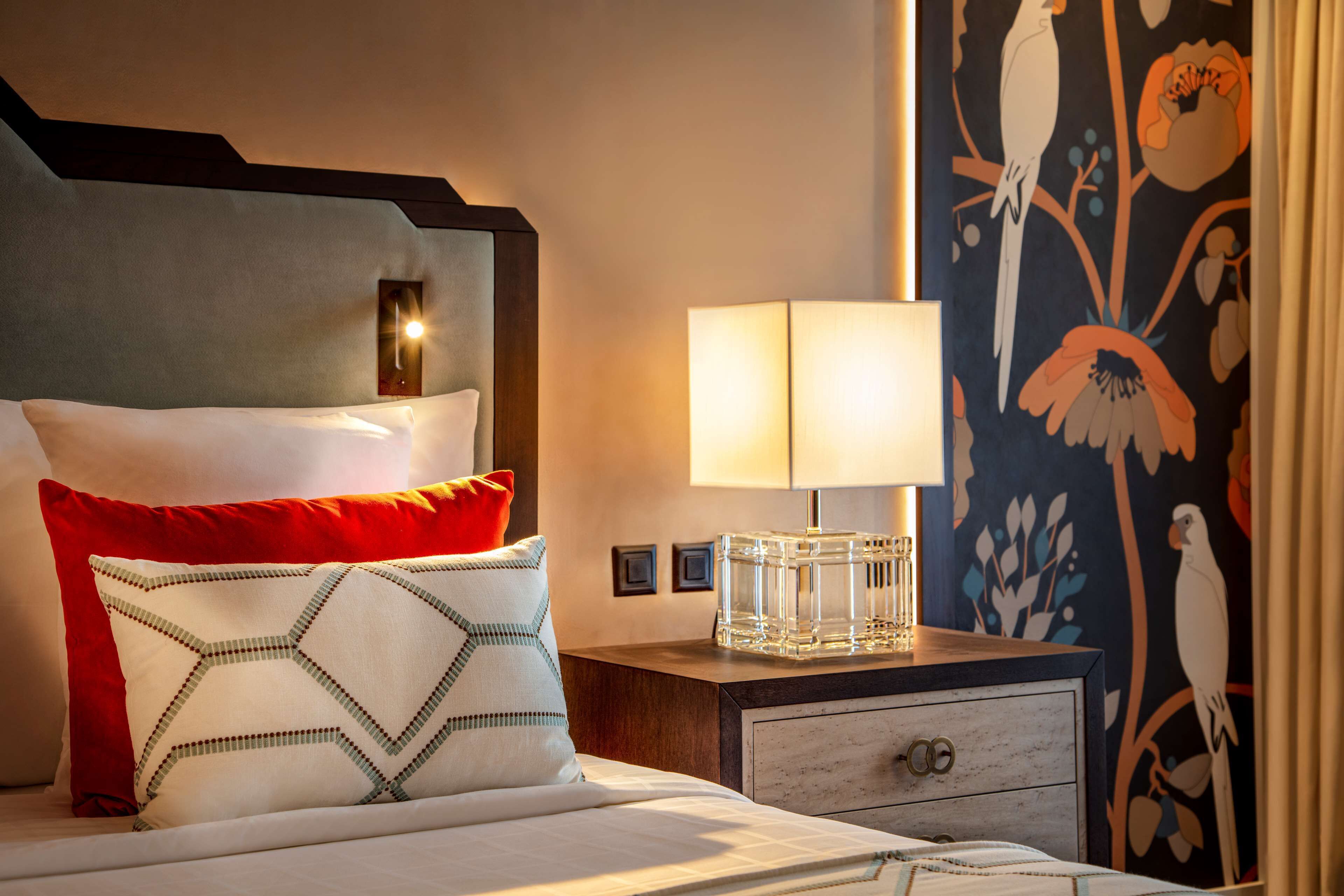 A neatly made bed with a red and white pillow, a clear glass vase on a wooden nightstand, and a wall with a bird mural.