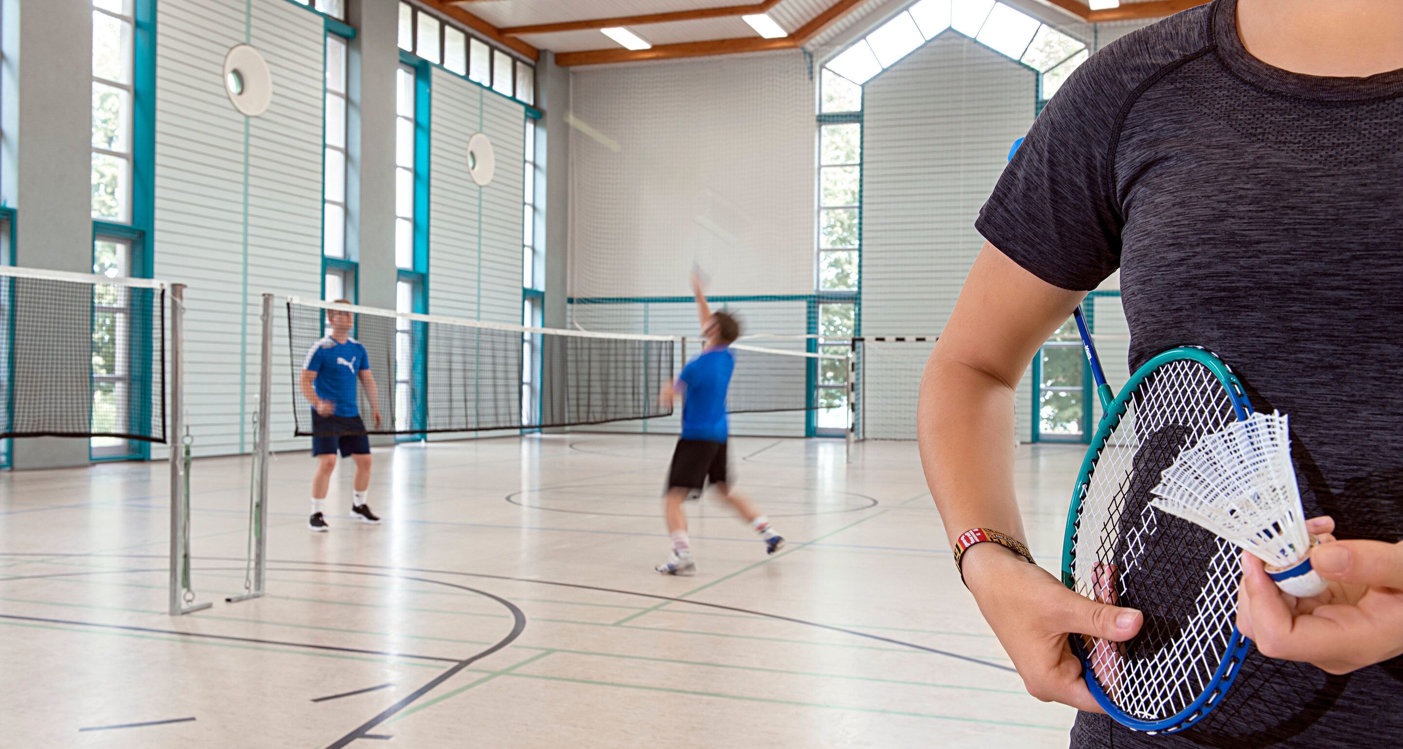 Ein Hallenbadmintonplatz mit Netz, Spielern und einer Person, die Badminton-Schlägern hält.