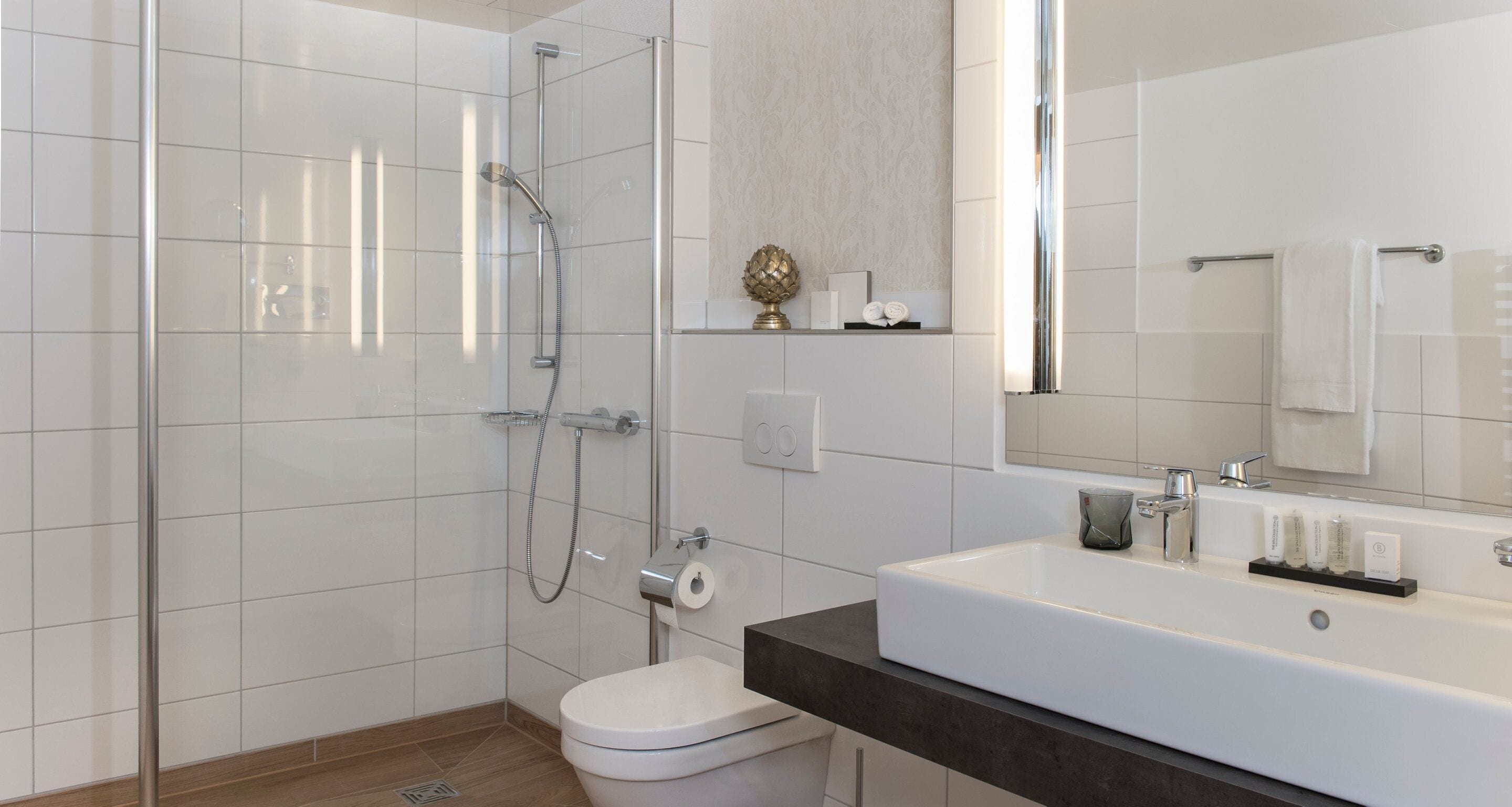 Un baño moderno con ducha envuelta en vidrio, inodoro blanco, espejo grande y encimera oscura con artículos de aseo.