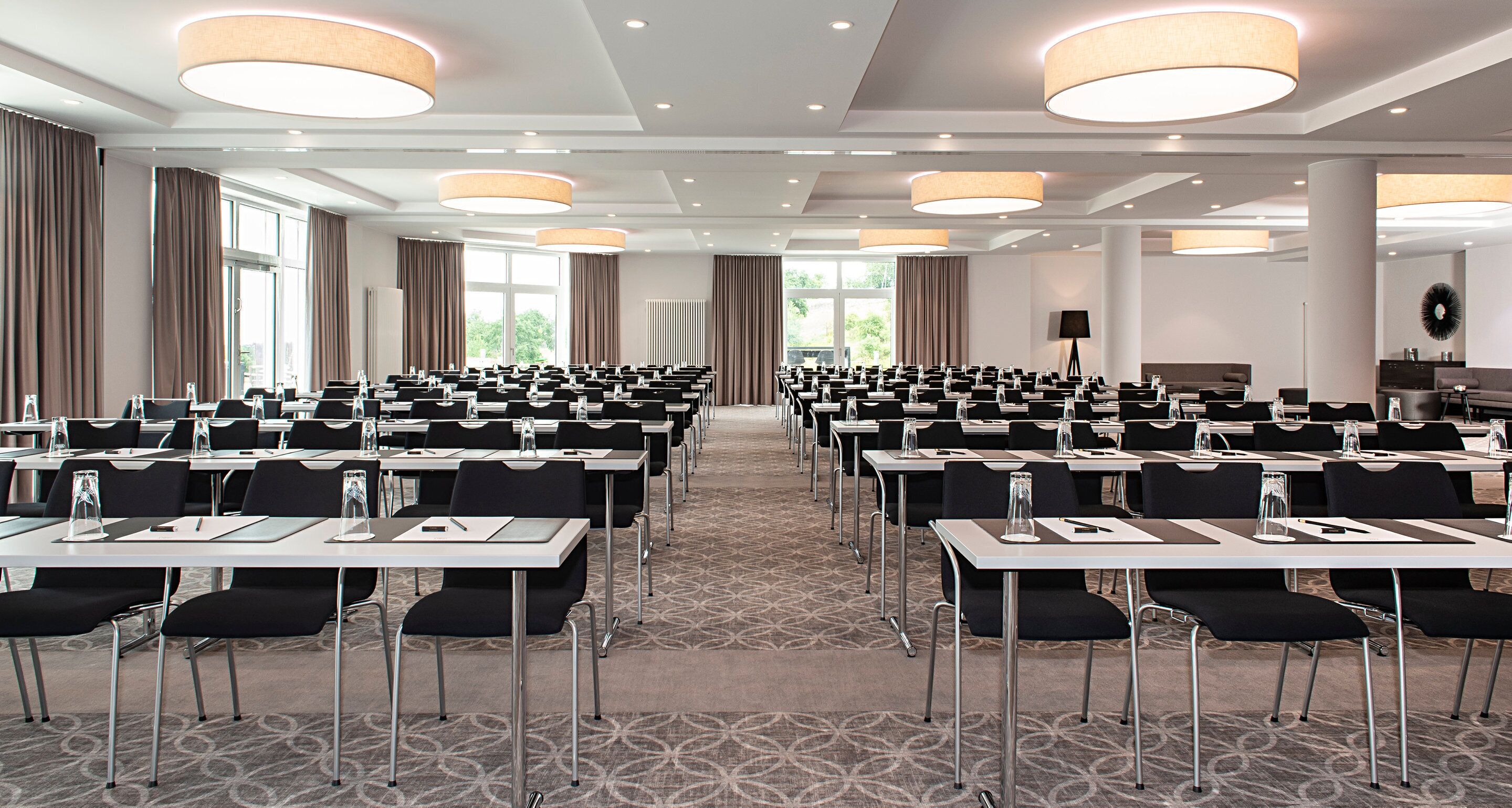 Une salle de conférence avec des rangées de tables et de chaises, de grandes fenêtres et des luminaires modernes.
