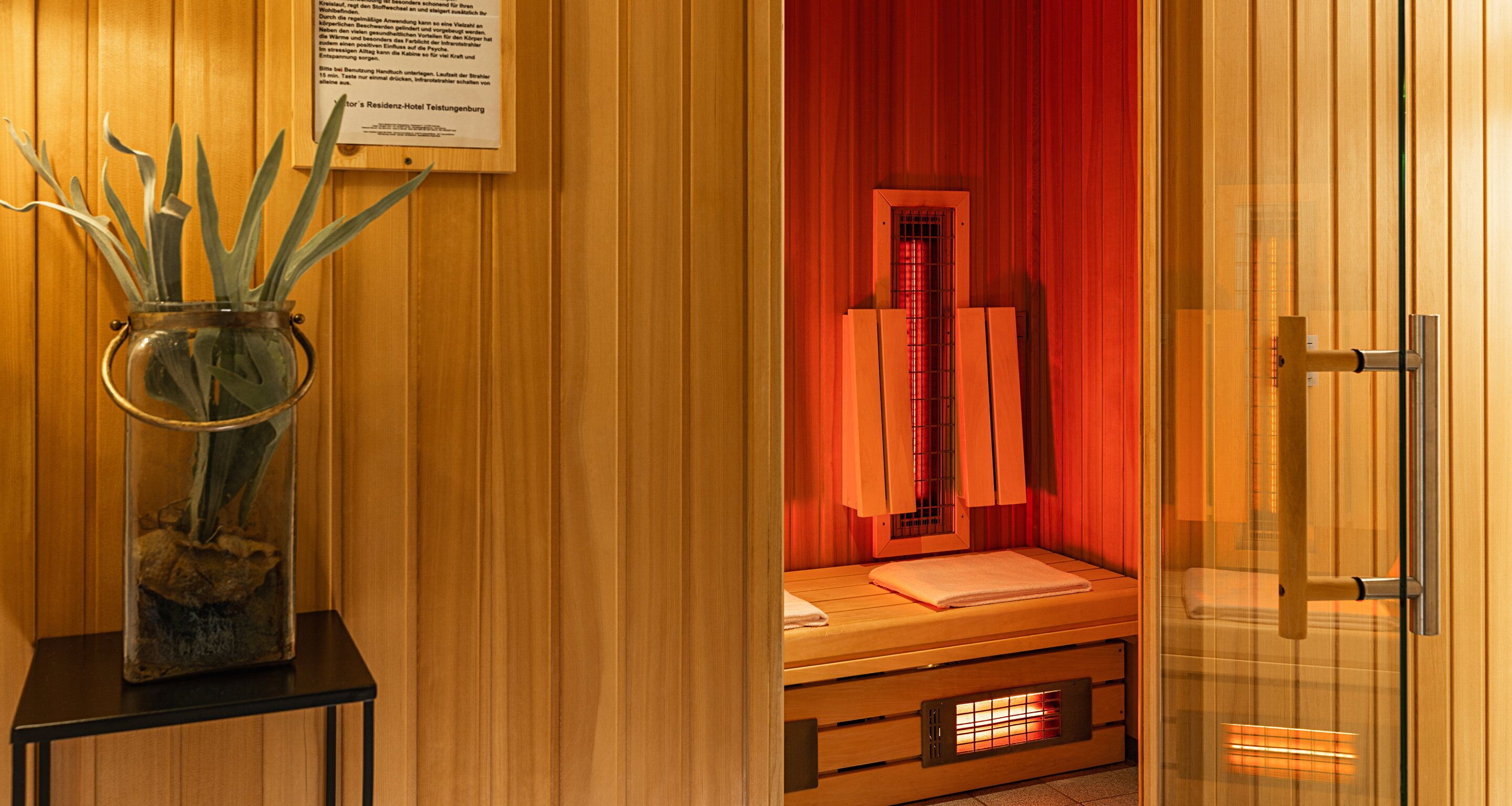 Una sala de sauna con un banco de madera, una puerta de cristal y un florero con plantas sobre una mesa.