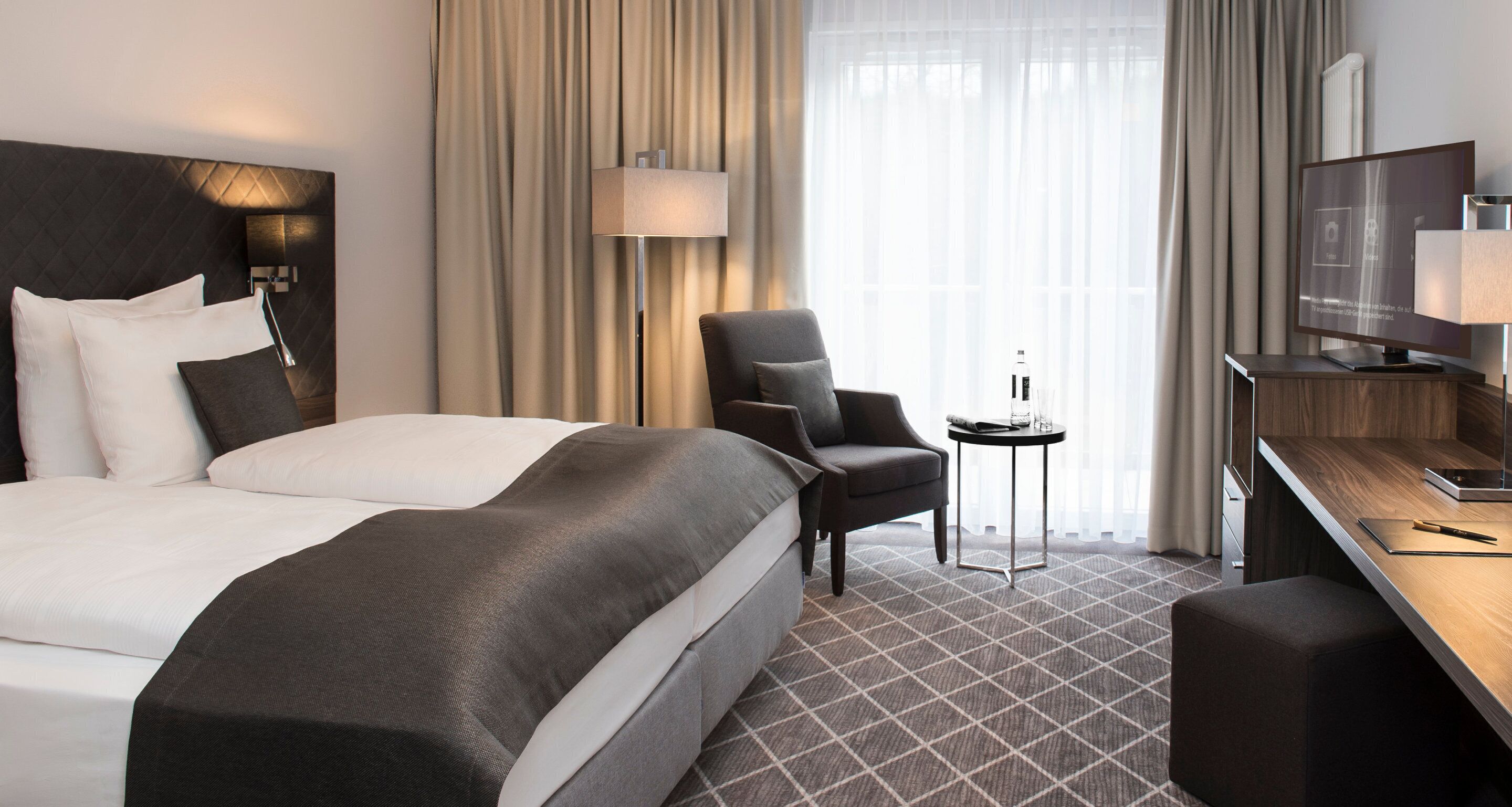A hotel room with a large bed, a desk, a chair, a small table, and a television.