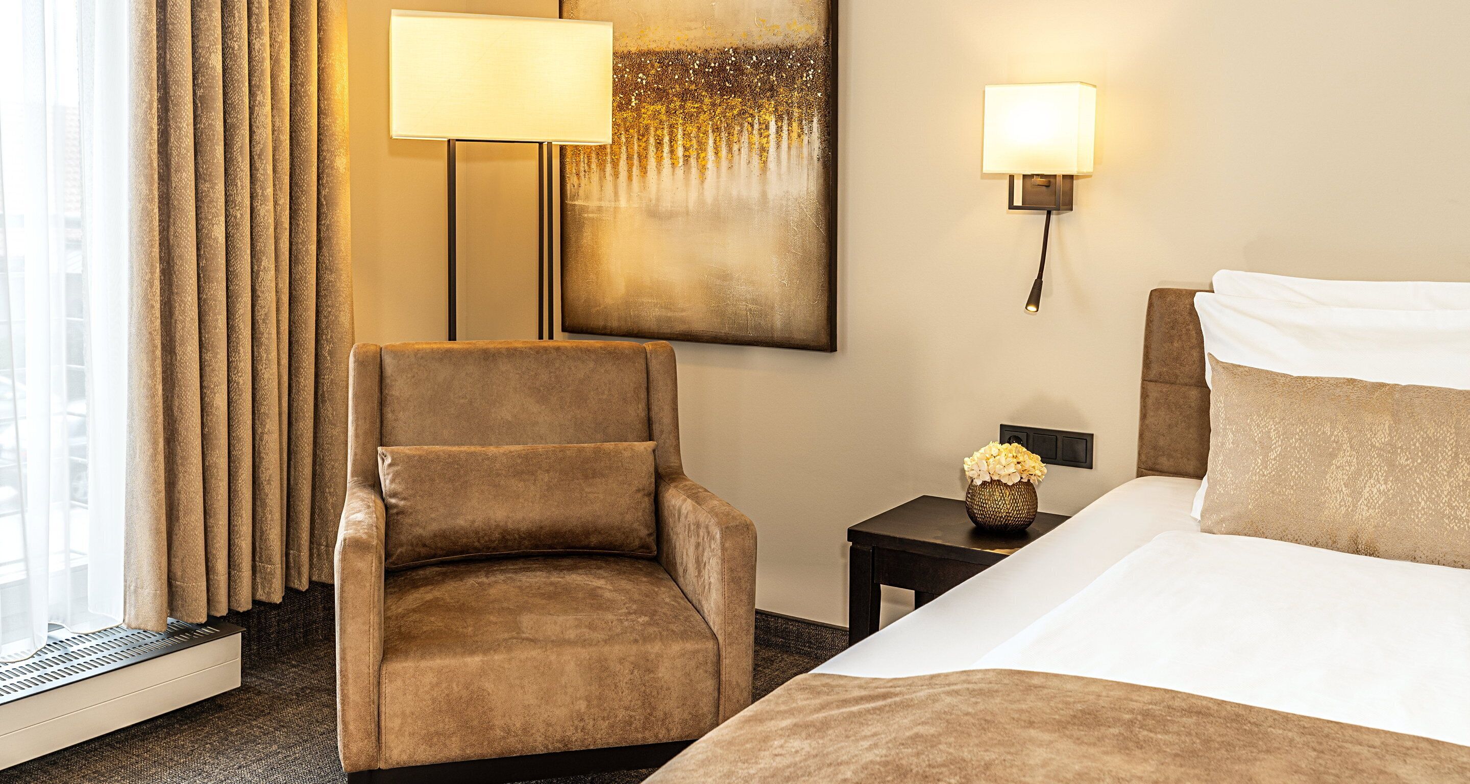 Habitación de hotel con una cama, una silla, una lámpara, un cuadro y una mesita de noche con un florero.