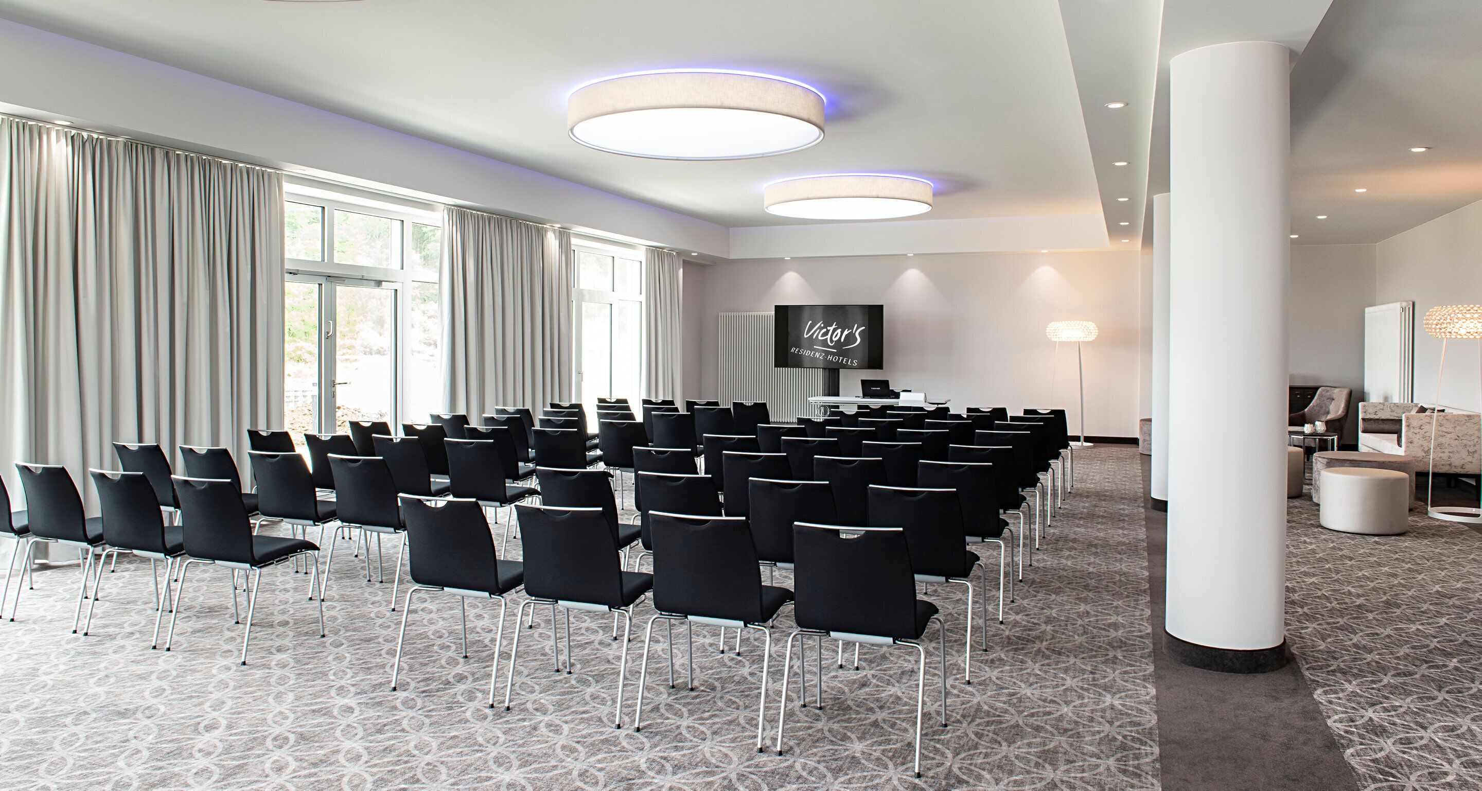 A conference room with rows of chairs, a large screen, and a reception area with a sofa and lamp.