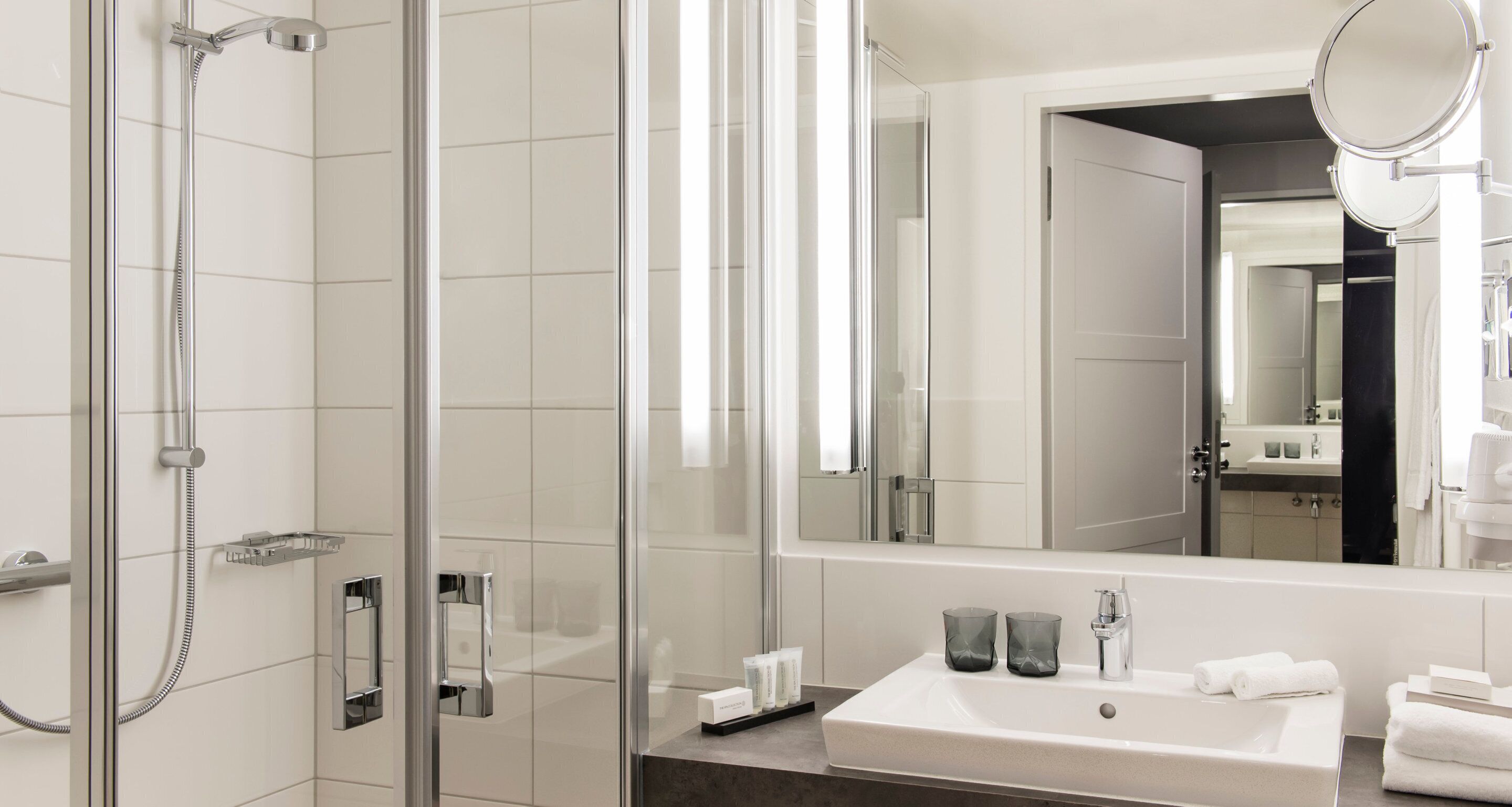 Un baño moderno con ducha envuelta en vidrio, un espejo grande y un lavamanos con artículos de aseo.