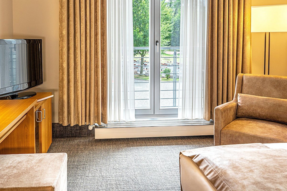 Una habitación de hotel con televisión, sofá, sillón, cama y ventana con cortinas.