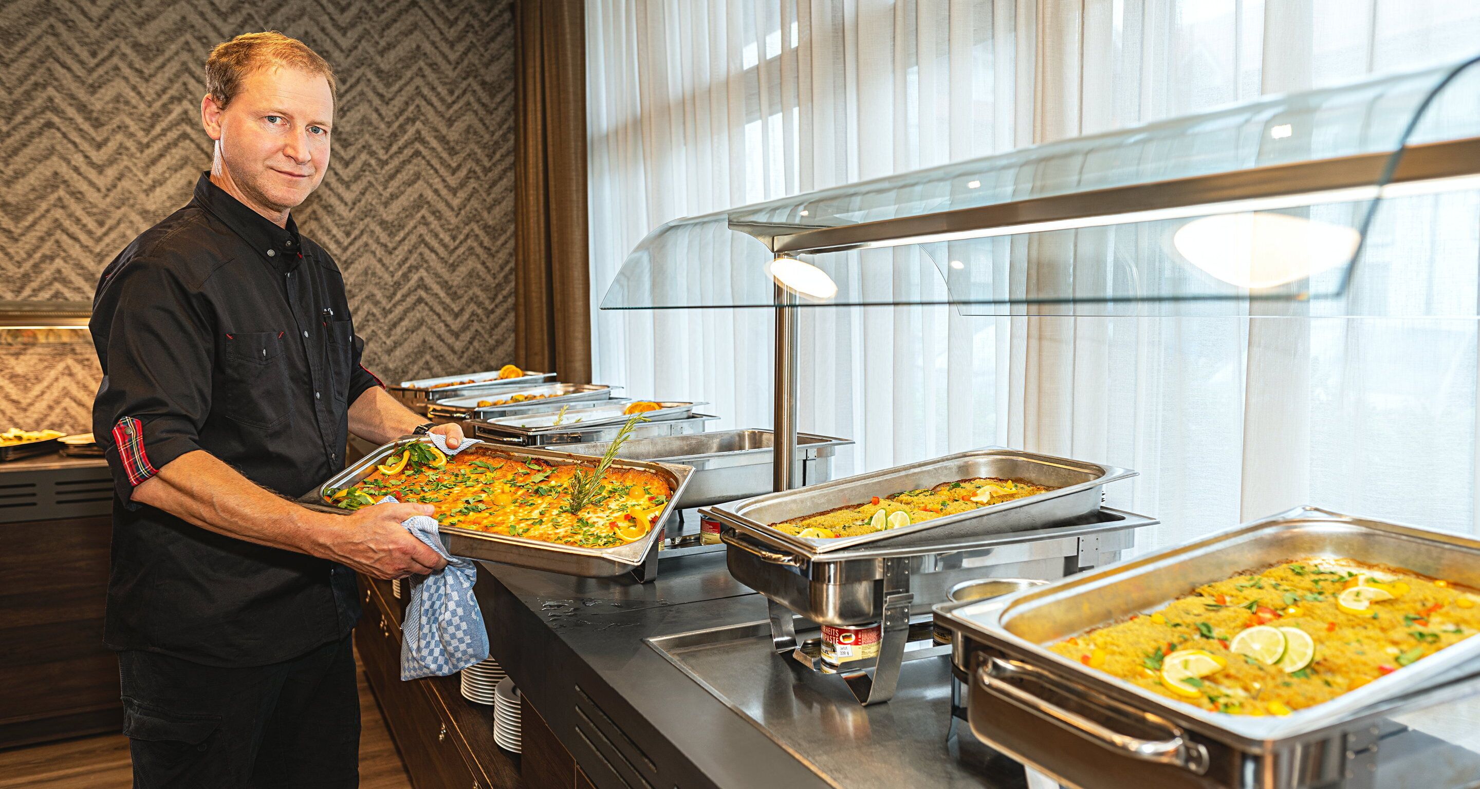 Ein reichhaltiges Buffet mit verschiedenen Speisen, eine Person bedient die Gäste, und ein modernes Ambiente mit gemustertem Wandpapier und großen Fenstern.