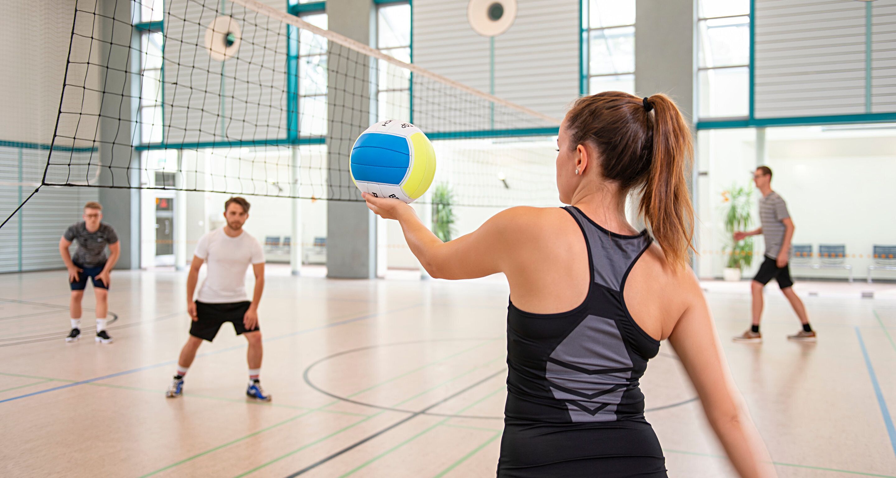 A volleyball game in an indoor gym with a high ceiling and large windows.