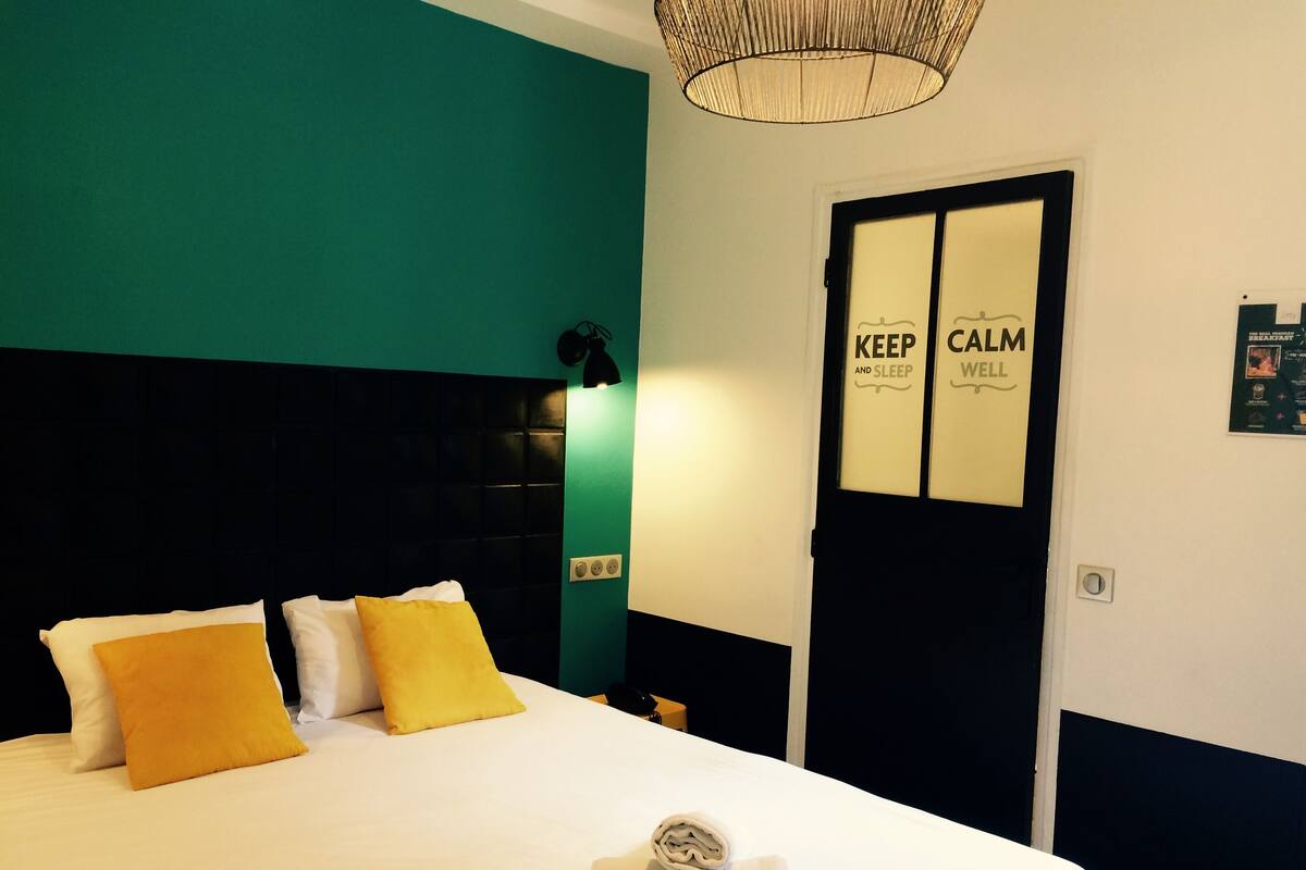 Ein ordentlich bezogenes Bett mit weißen und gelben Kissen, ein gefaltetes Handtuch und ein schwarzes Kopfteil. Eine Tür mit dem Schriftzug "KEEP CALM AND SLEEP" ist sichtbar.