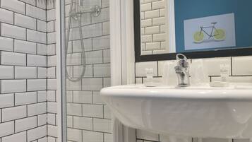 Une salle de bain avec une douche à parois de verre, des carreaux de céramique blancs rectangulaires, un lavabo blanc et un miroir orné d’un cadre avec une image.