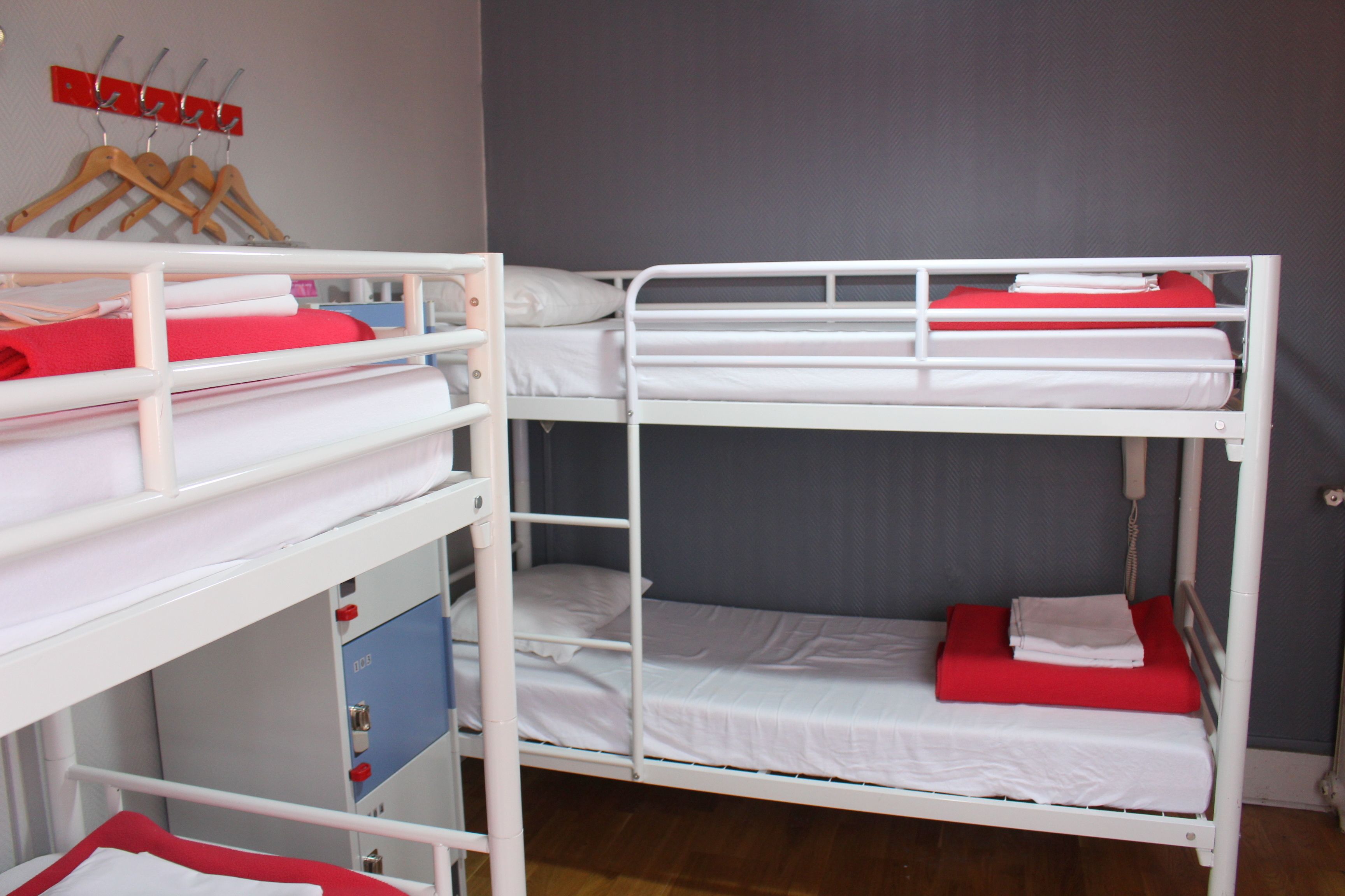 Une chambre avec un lit superposé, composé de deux lits, chacun étant équipé de draps blancs et d’oreillers rouges. Un porte-vêtements en bois, sur lequel sont accrochés des vêtements, est fixé au mur au-dessus des lits.