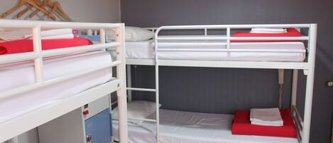 Une chambre avec un lit superposé, composé de deux lits, chacun étant équipé de draps blancs et d’oreillers rouges. Un porte-vêtements en bois, sur lequel sont accrochés des vêtements, est fixé au mur au-dessus des lits.