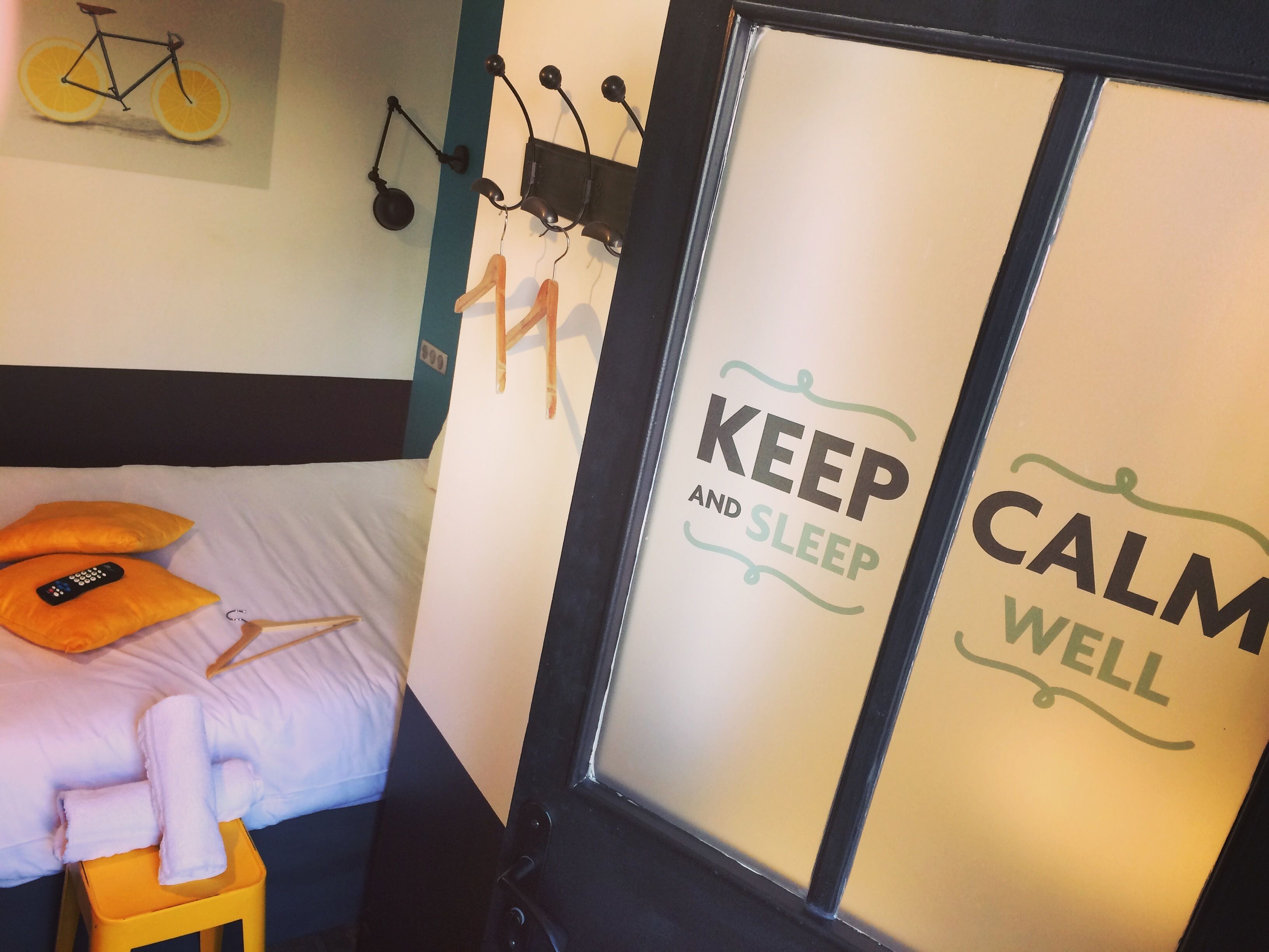 A hotel room with a bed, a small stool with towels, a wall-mounted bicycle, and a door with a motivational sign.