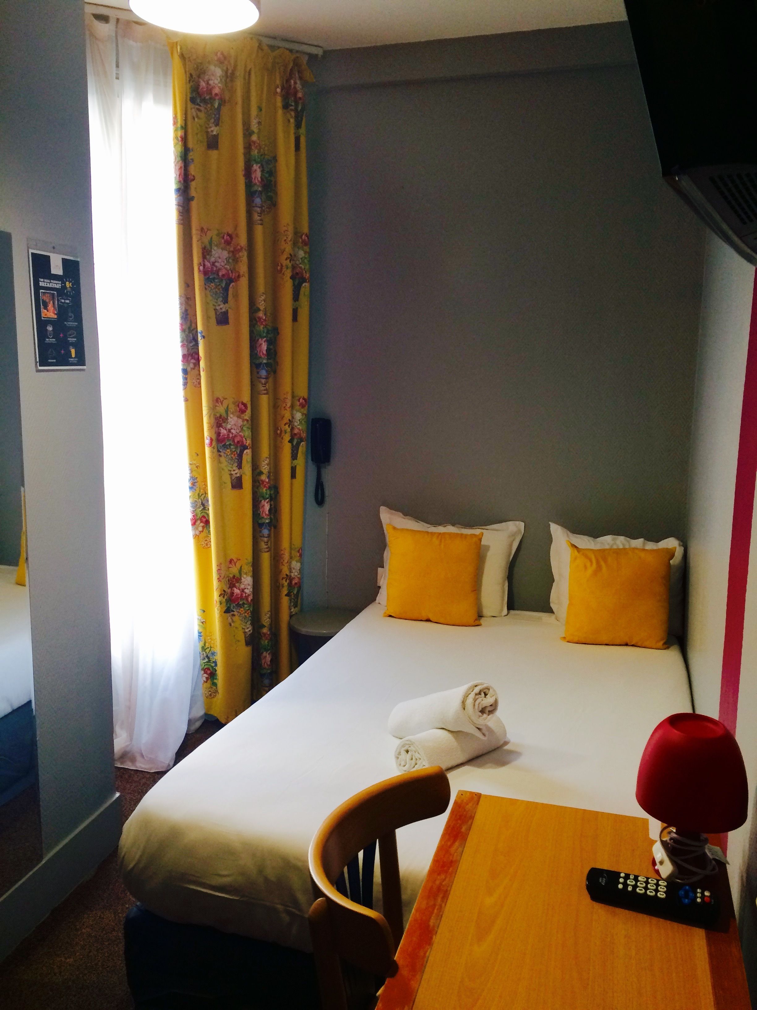A hotel room with a bed, two pillows, a small table with a remote control, and a chair.