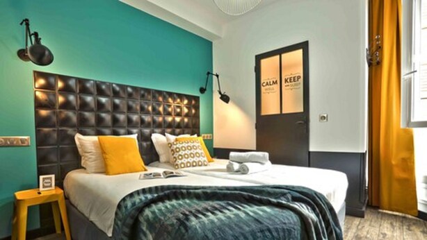 A hotel room with a tufted headboard, a bed with a blue and white duvet, a small wooden nightstand, a wall-mounted lamp, and a door with a 'CALM KEEP' sign. - Arty Paris Porte de Versailles by River - Hostel (Paris)