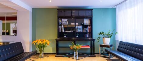 A modern lobby with a black leather sofa, a glass table with a vase of flowers, and a bookshelf against a teal wall.