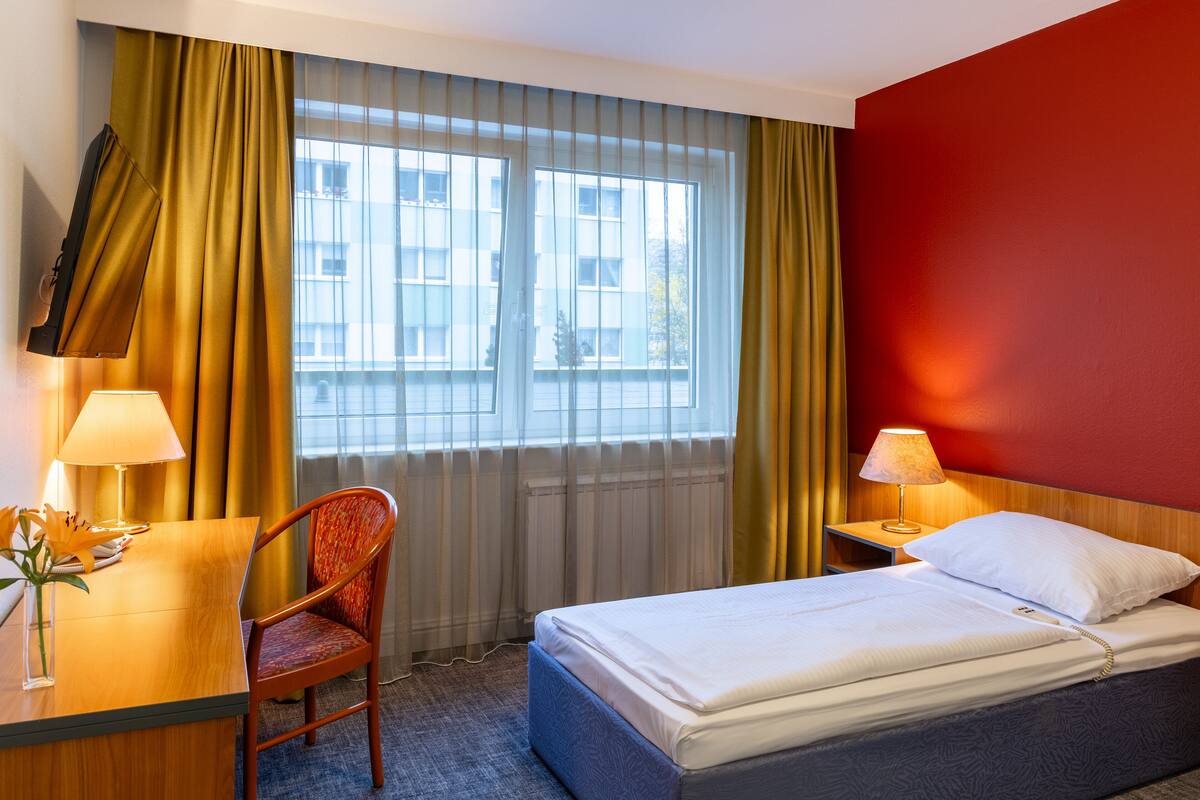 Een hotelkamer met een bed, een bureau met lamp, een stoel en uitzicht op gebouwen door het raam.