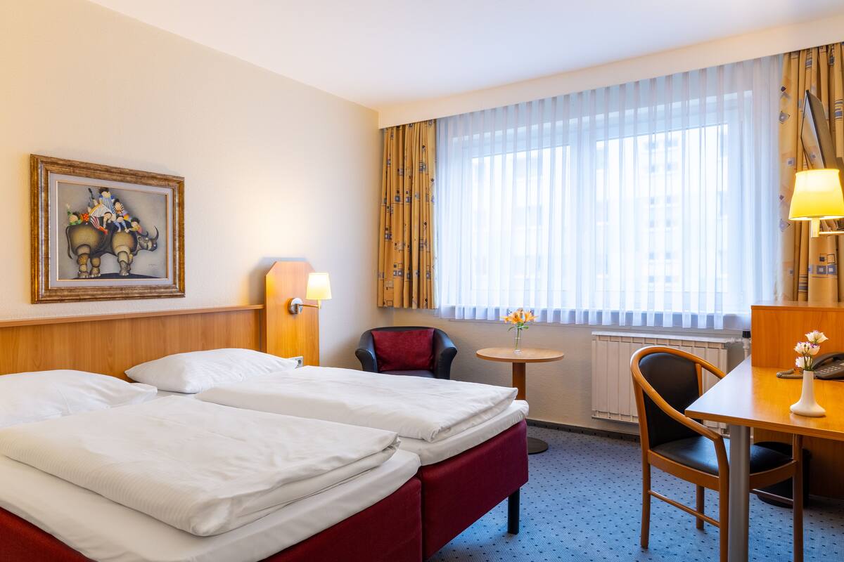 Een hotelkamer met twee bedden, een bureau, een stoel, een lamp, een ingelijste foto aan de muur en een raam met gordijnen.