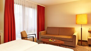 Una habitación de hotel con cama, sofá, sillón, una mesita con fruta y una lámpara.