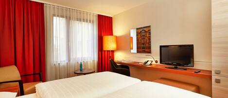 A hotel room with a large bed, a desk with a television, a chair, a lamp, and a window with red curtains.
