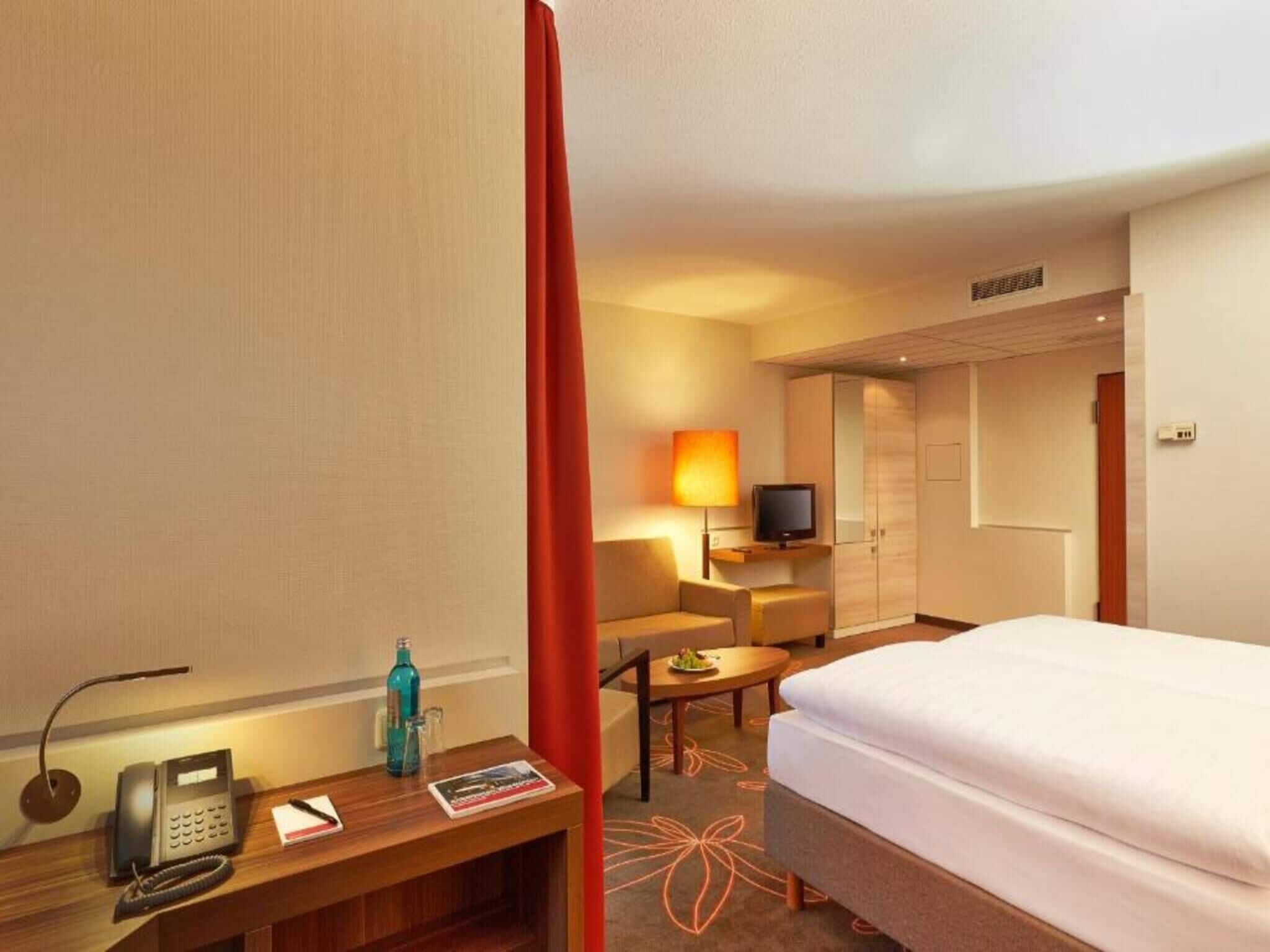 A hotel room with a bed, a desk with a telephone, a television, and a small table with a lamp.