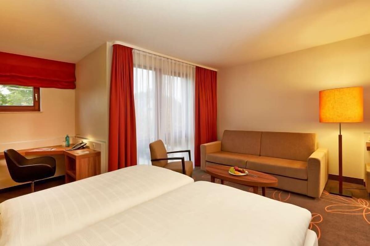 A hotel room with a bed, a desk, a chair, a sofa, and a lamp.