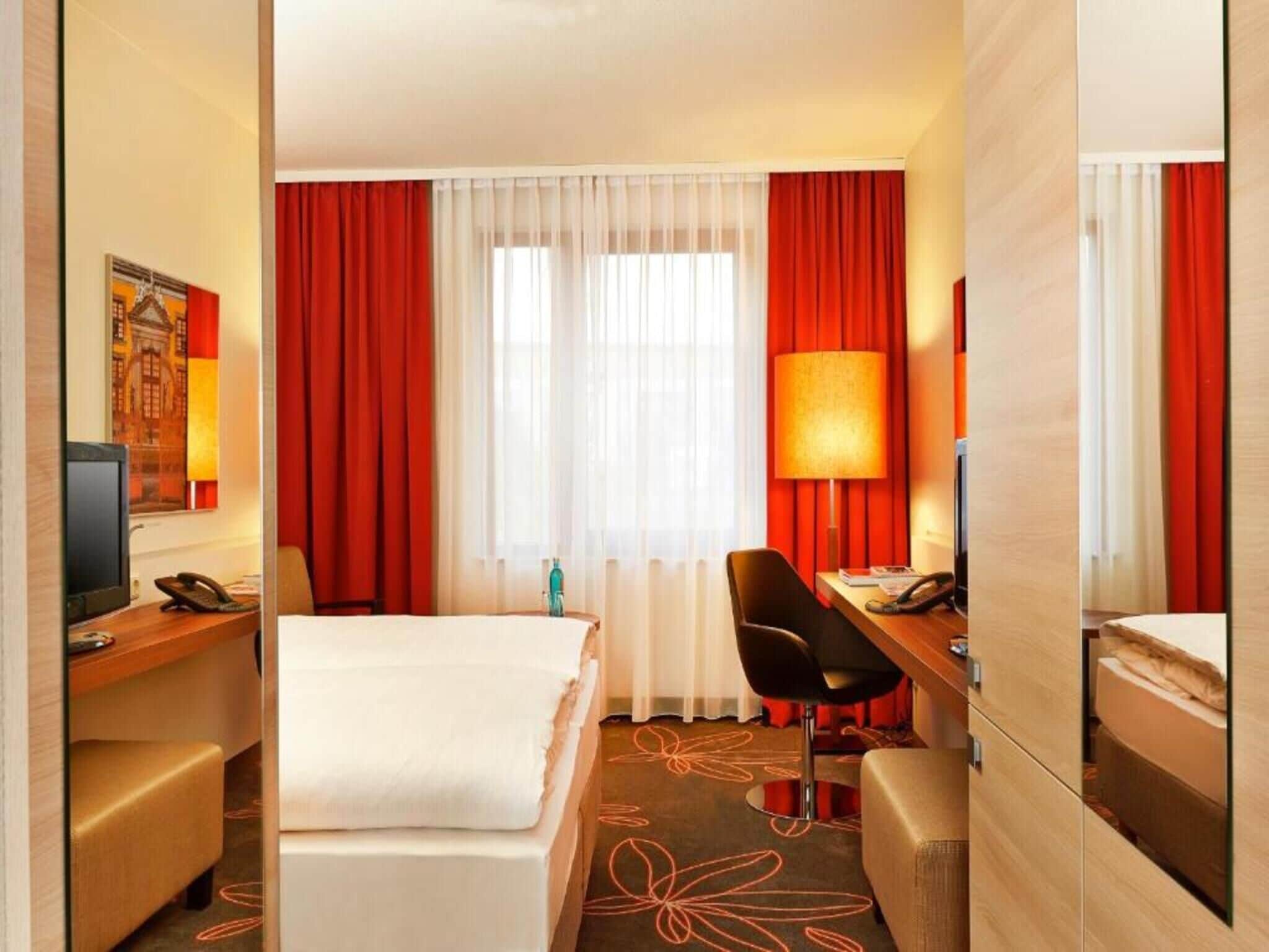 Habitación de hotel con cama, escritorio, silla, televisor y ventana con cortinas.