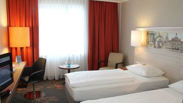 Ein Hotelzimmer mit zwei Betten, einem Schreibtisch mit Computer, einem Stuhl, einem kleinen Tisch mit Lampe und einem Fenster mit Vorhängen.