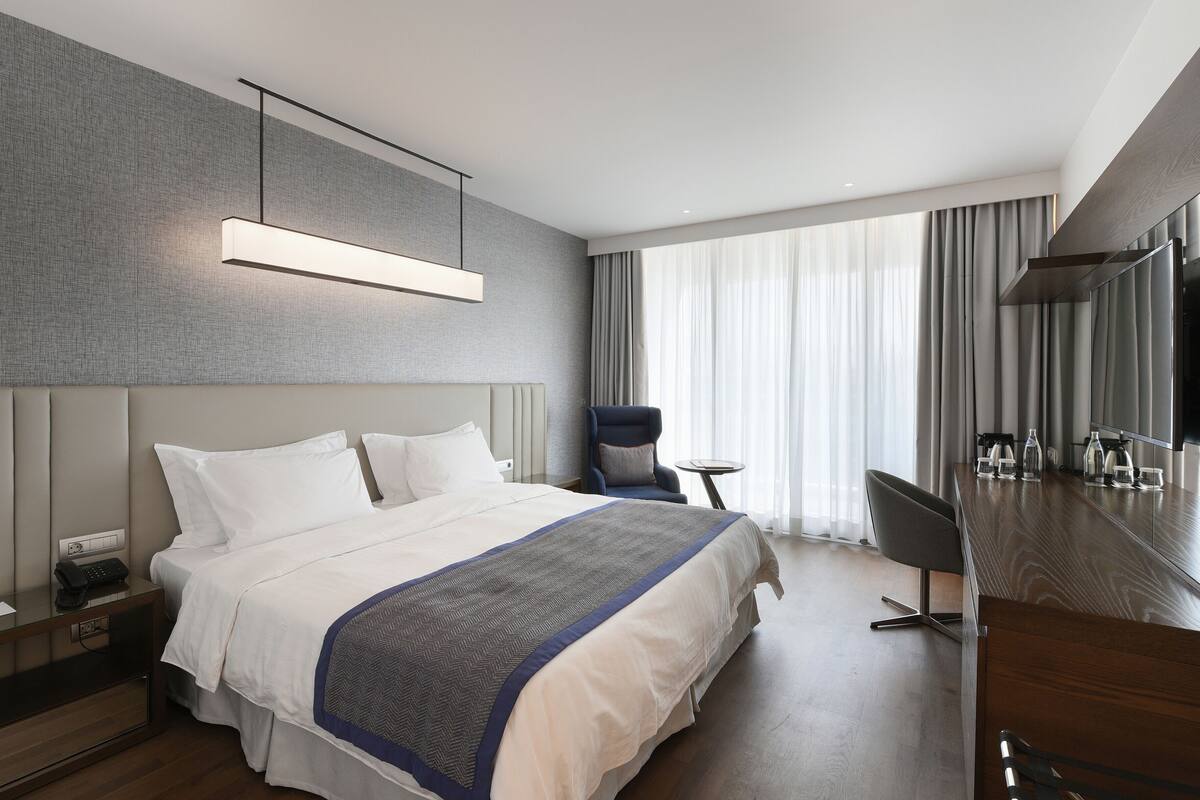 A modern hotel room with a large bed, a desk with a chair, a small table, and a TV.