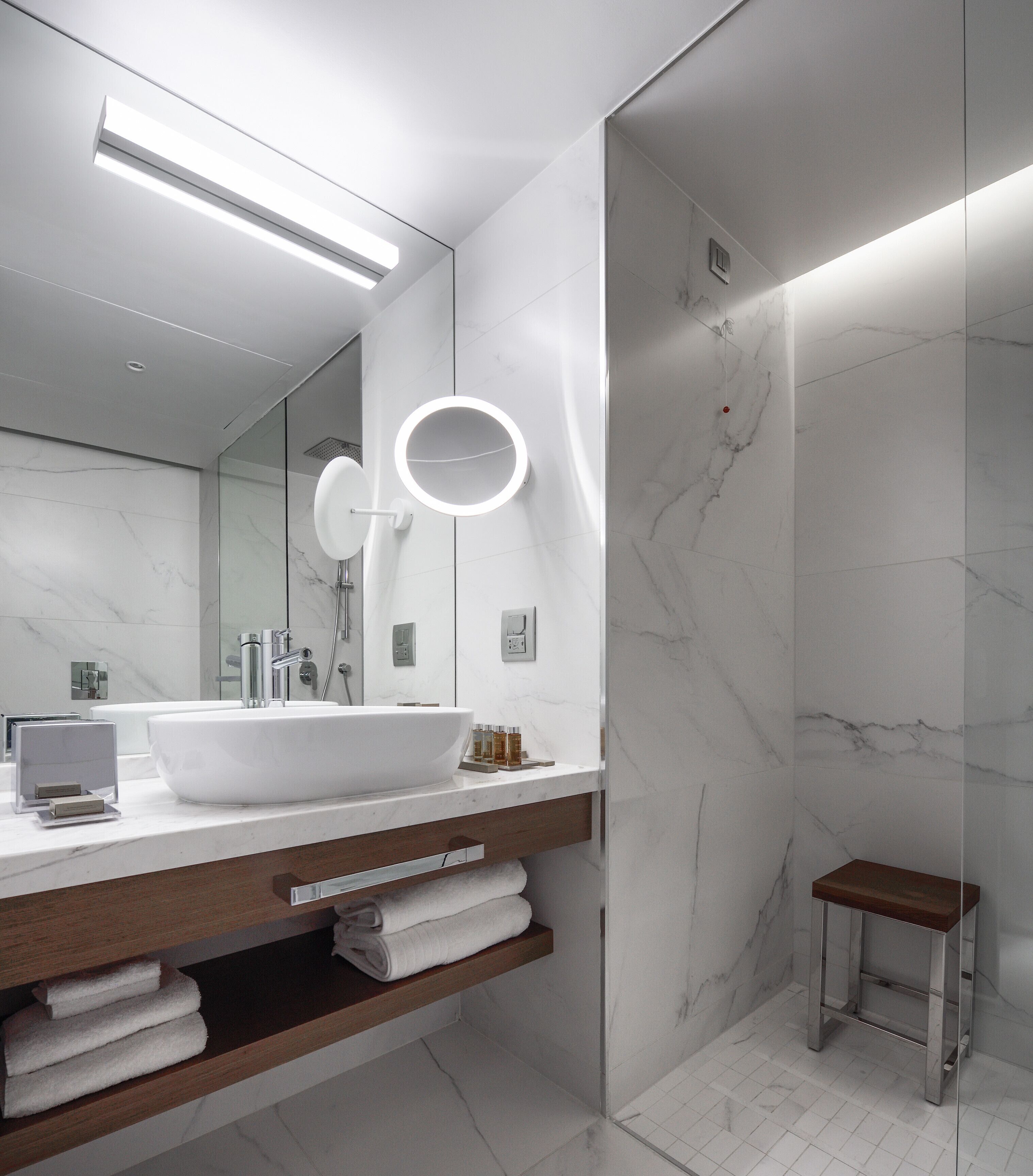 Un baño moderno con un lavamanos grande de color blanco, un espejo con iluminación integrada, una zona de ducha y un taburete pequeño de madera.