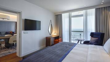A hotel room with a bed, a desk, a chair, a TV, and a balcony with a view of the sea.