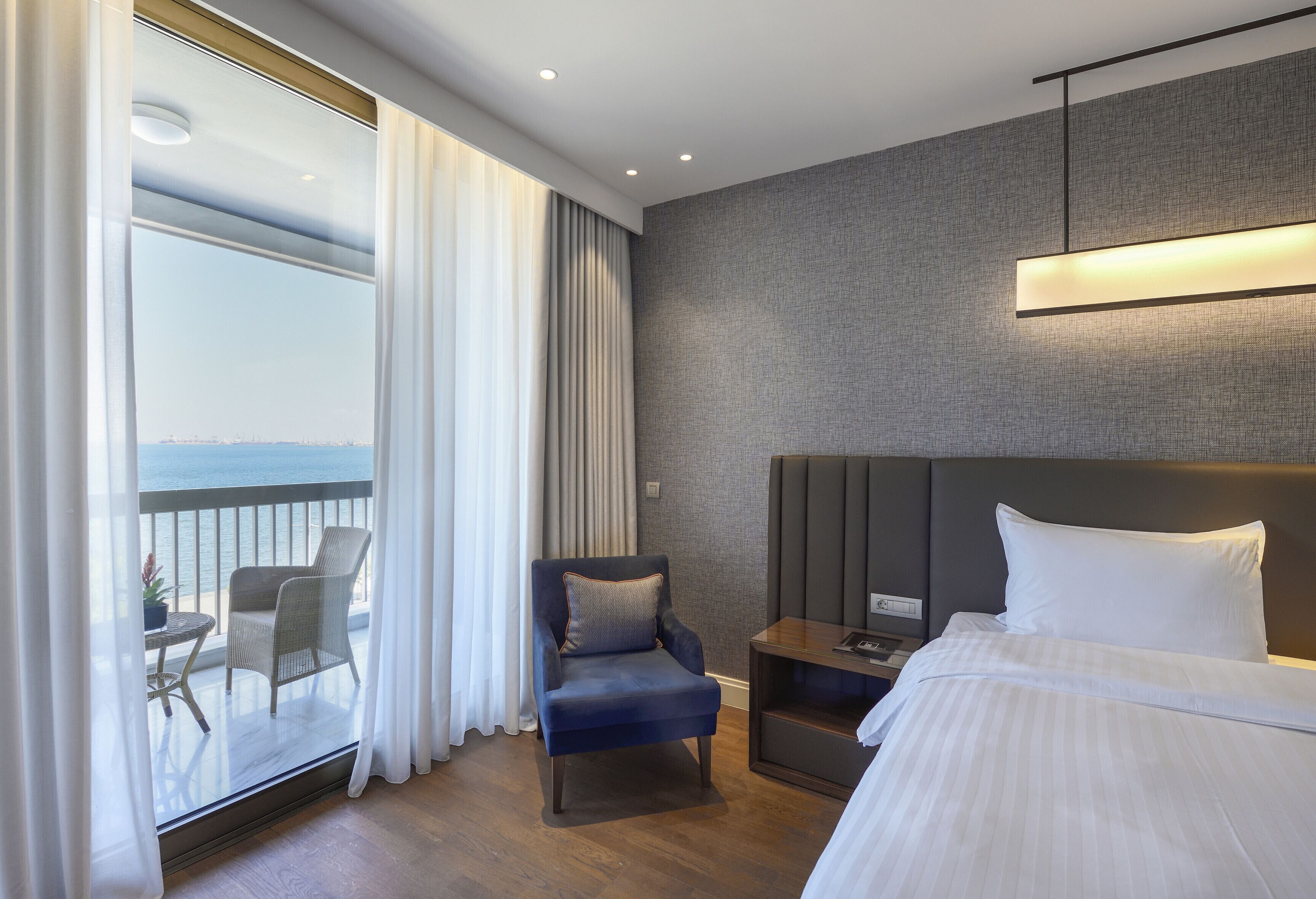 Habitación de hotel con cama, una silla, una mesita pequeña, una mesita de noche y vistas al océano a través de amplias ventanas.
