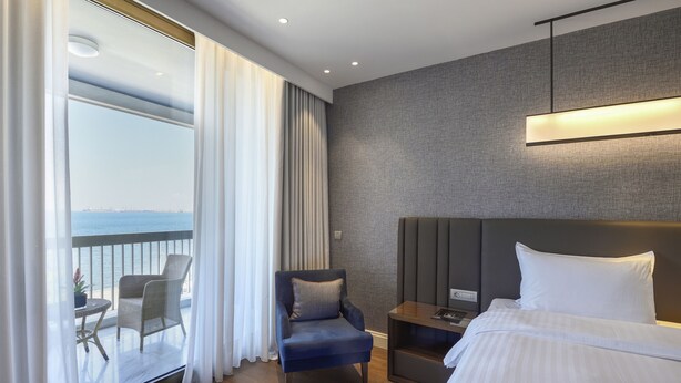 A hotel room with a bed, a chair, a small table, a nightstand, and a view of the ocean through large windows. - Makedonia Palace (Thessaloniki)