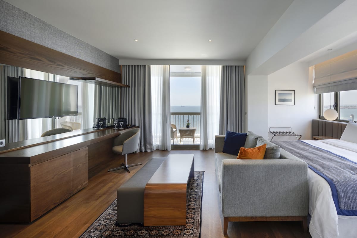 A modern hotel room with a large bed, a sofa, a desk with a TV, and a balcony with a view of the sea.