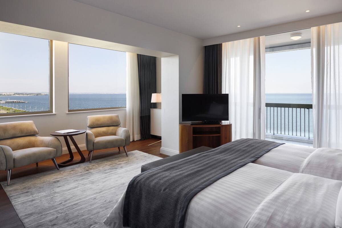 A hotel room with a large bed, a TV, a seating area with a chair and table, and a view of the sea.