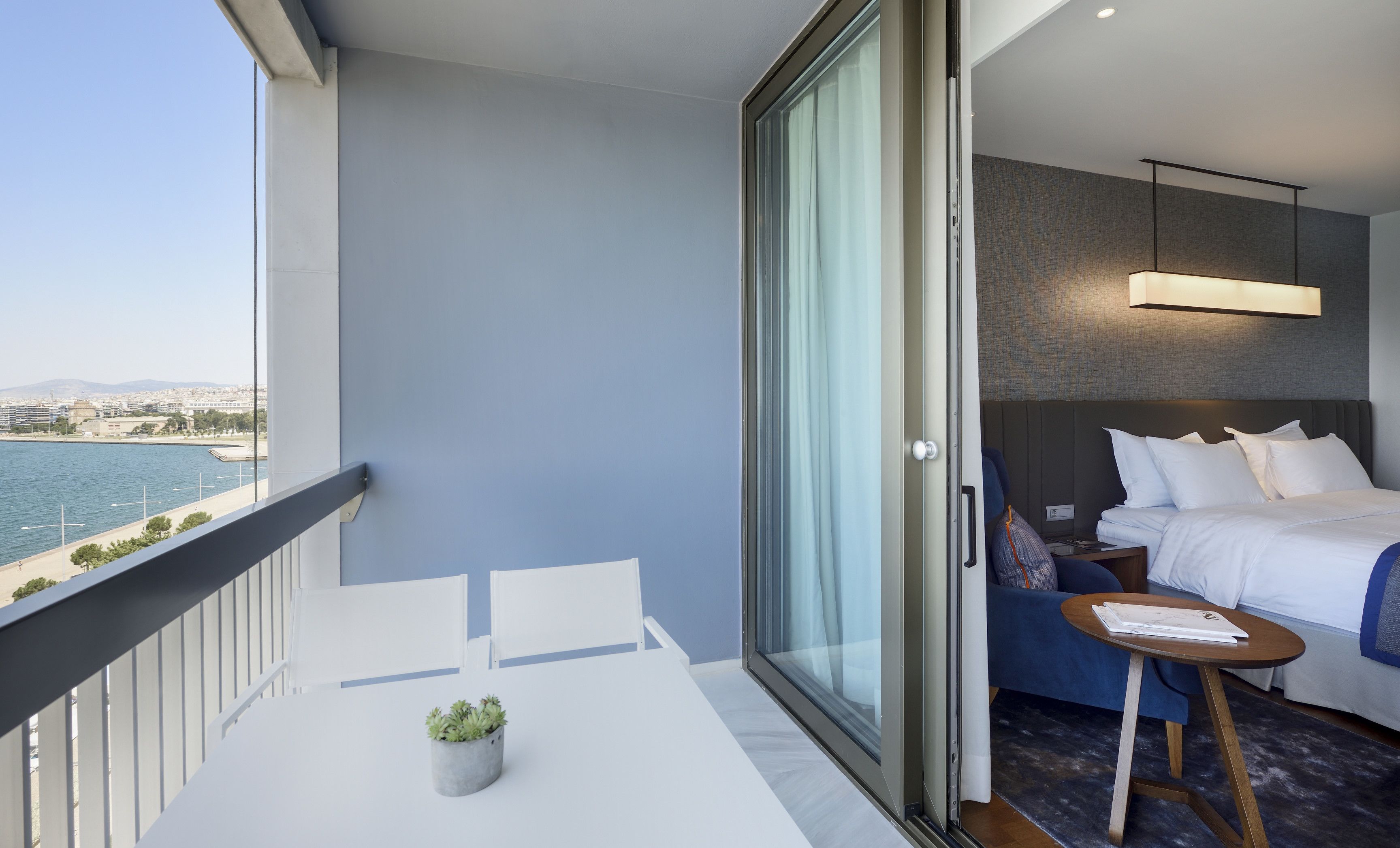 A modern hotel room with a balcony overlooking a body of water, a bed with white linens, and a small table with a plant.