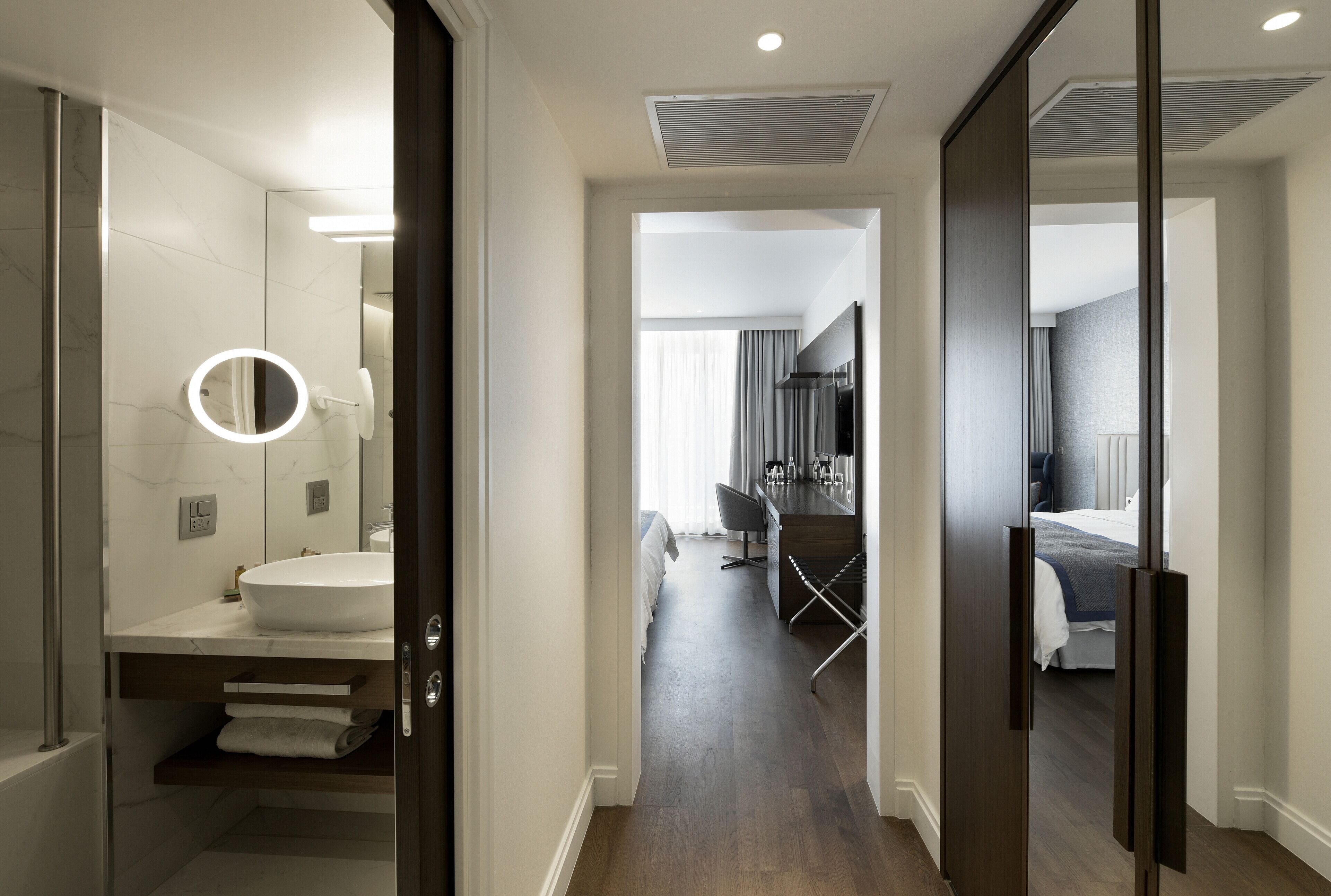 Une chambre d’hôtel moderne dotée d’une salle de bain comprenant un grand miroir, un lavabo et un porte-serviettes. La chambre a un plancher en bois et un lit avec une tête de lit.