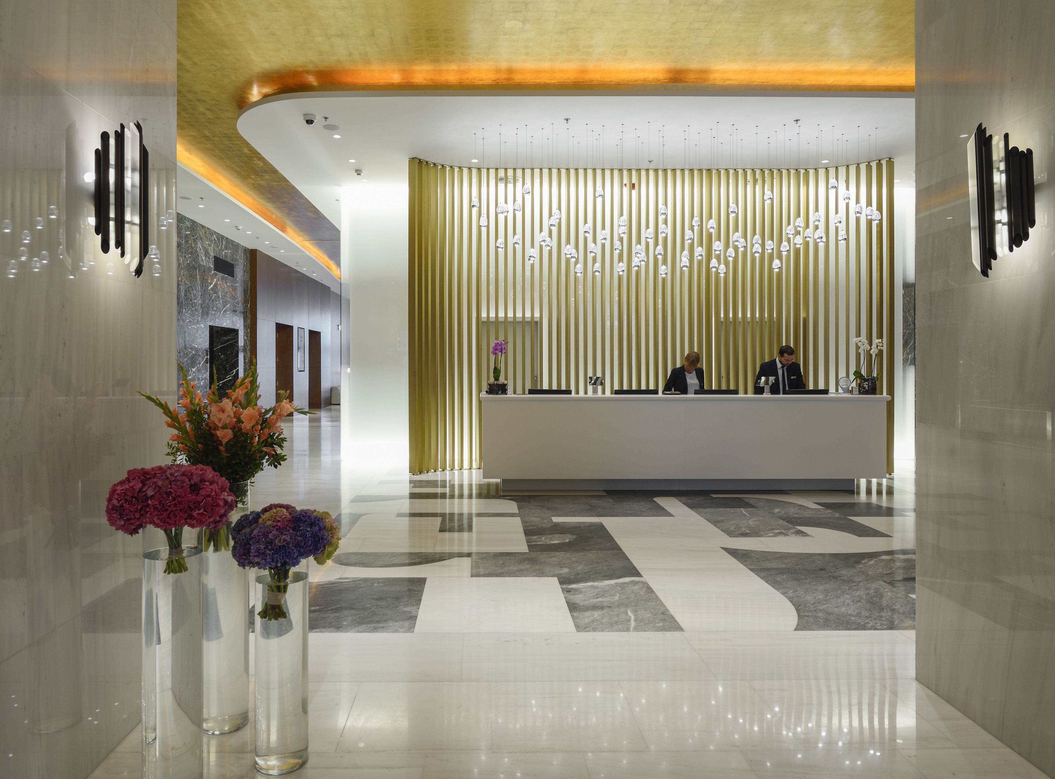 A modern hotel lobby with a reception desk, a decorative wall with hanging lights, and a marble floor.