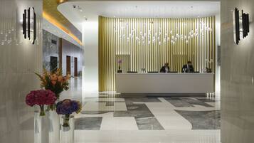 A modern hotel lobby with a reception desk, a decorative wall with hanging lights, and a marble floor.