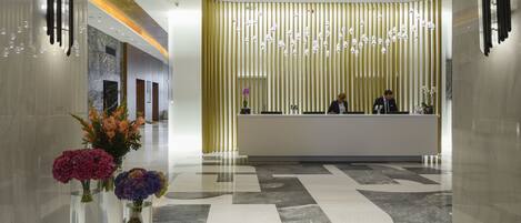 A modern hotel lobby with a reception desk, a decorative wall with hanging lights, and a marble floor.