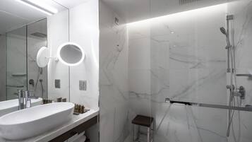 Un baño moderno con un lavamanos grande blanco, un espejo con luz circular, una zona de ducha y un taburete pequeño.