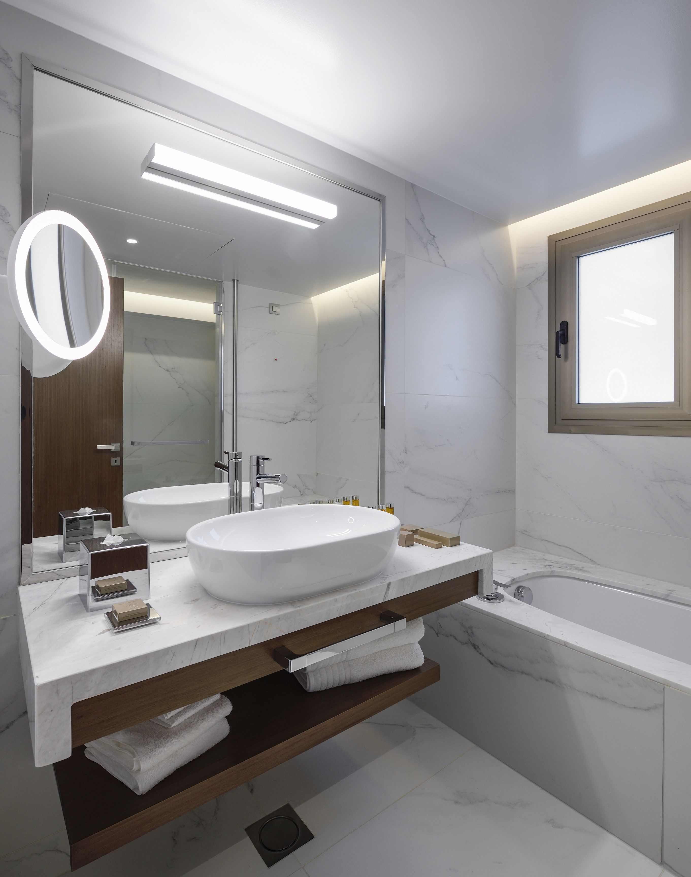 A modern bathroom with a large mirror, two sinks, a bathtub, and a window.
