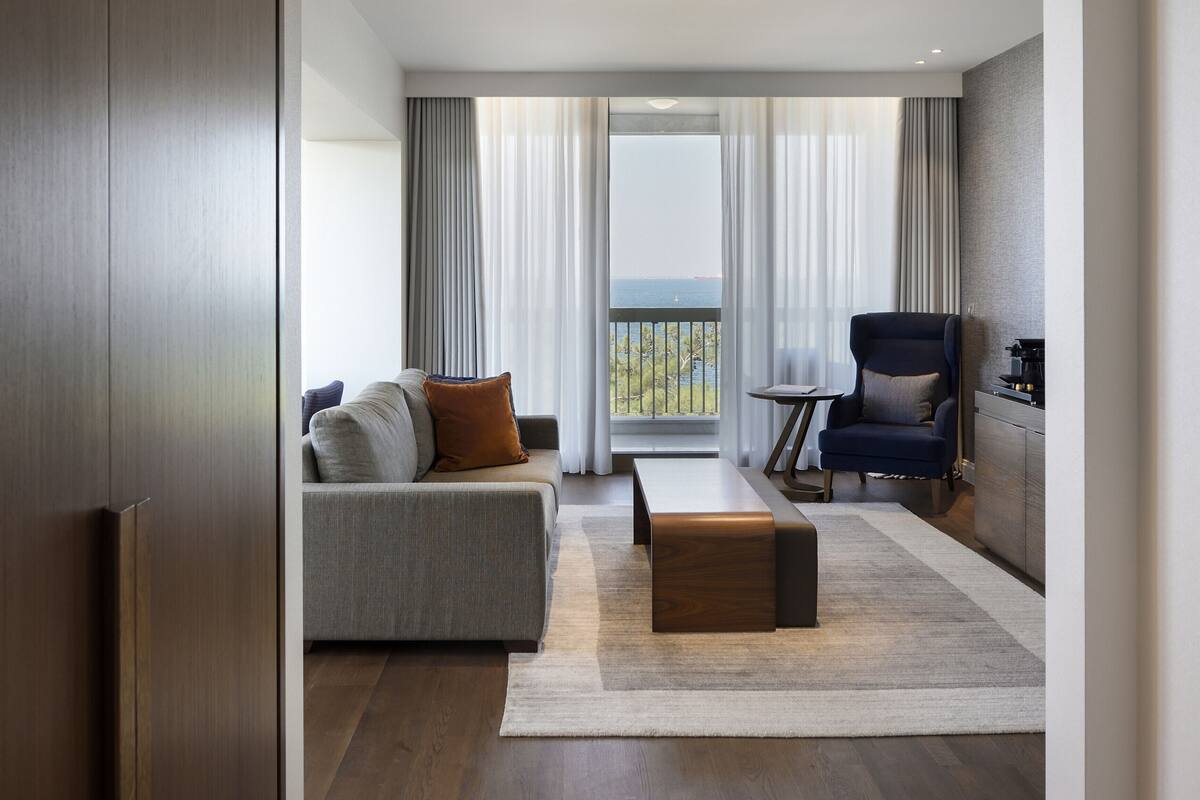 A modern living room with a sofa, a coffee table, and a chair. There is a view of the sea through large windows.