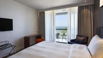 A hotel room with a large bed, a TV mounted on the wall, a small seating area with a chair and table, and a balcony with a view of the sea.