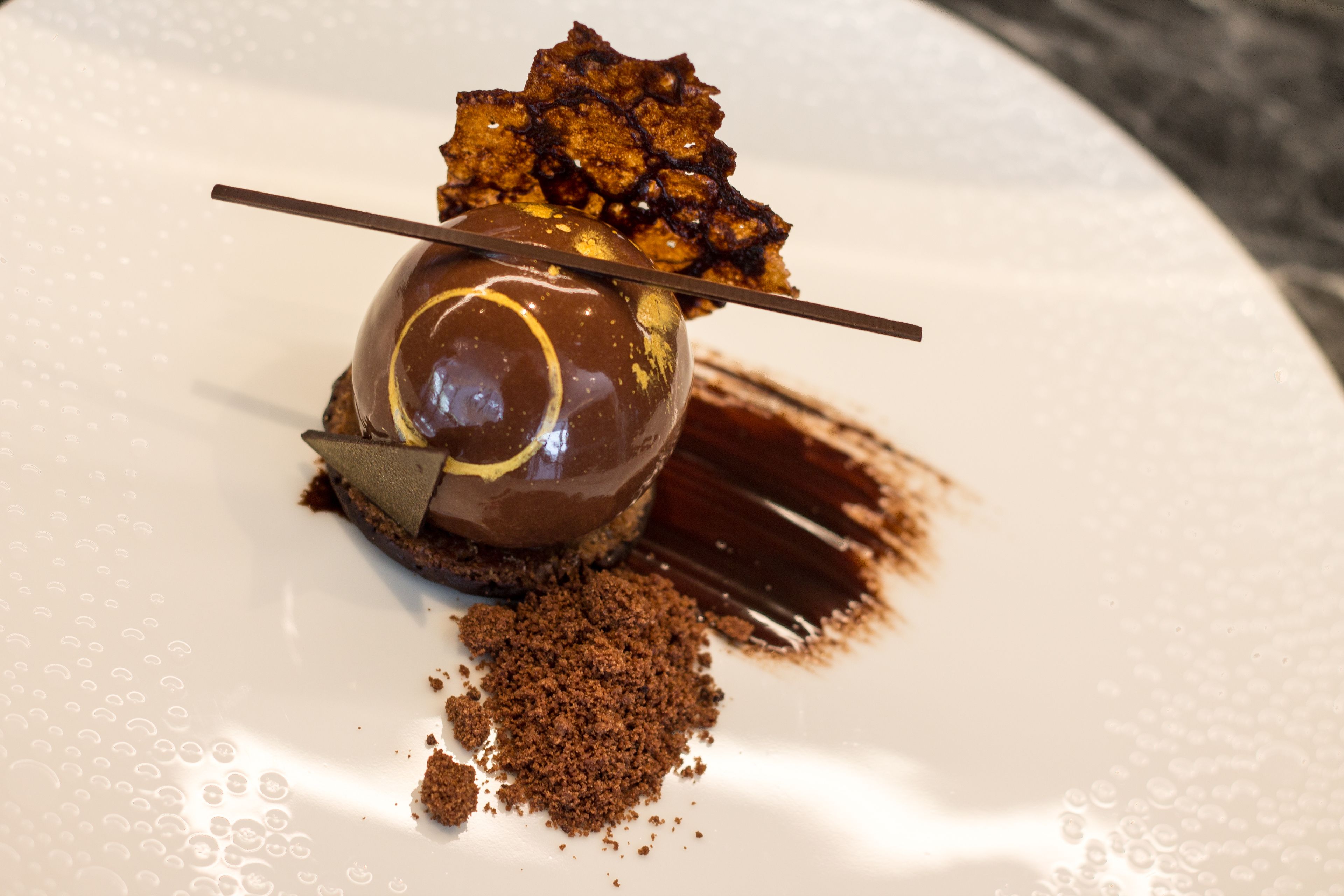 Postre sobre un plat blanc amb una esfera de xocolata, una cobertura caramel·litzada i un palinet de xocolata.
