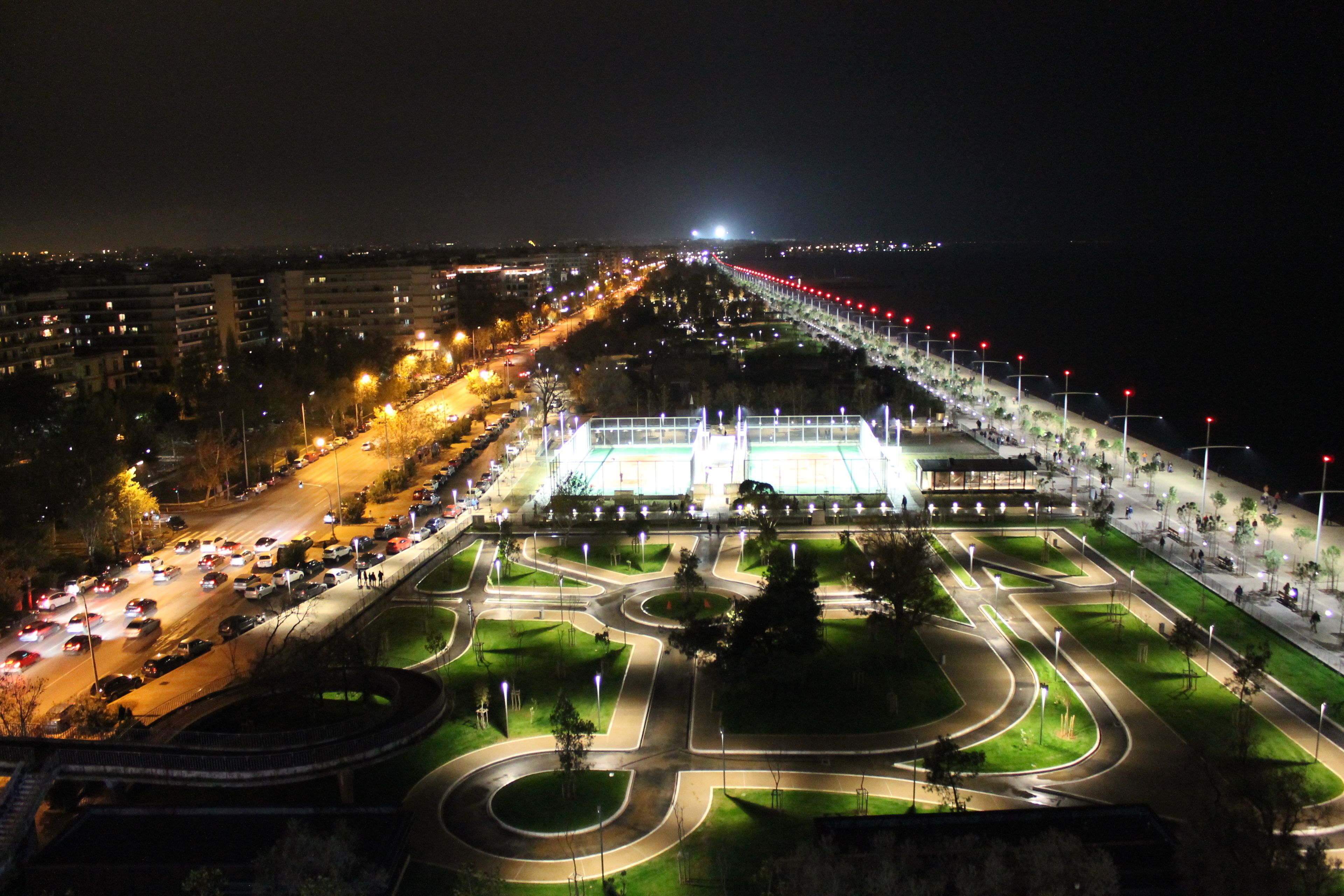 Vista aérea de un paisaje urbano nocturno con una glorieta bien iluminada, un edificio grande con techo verde y un río al fondo.